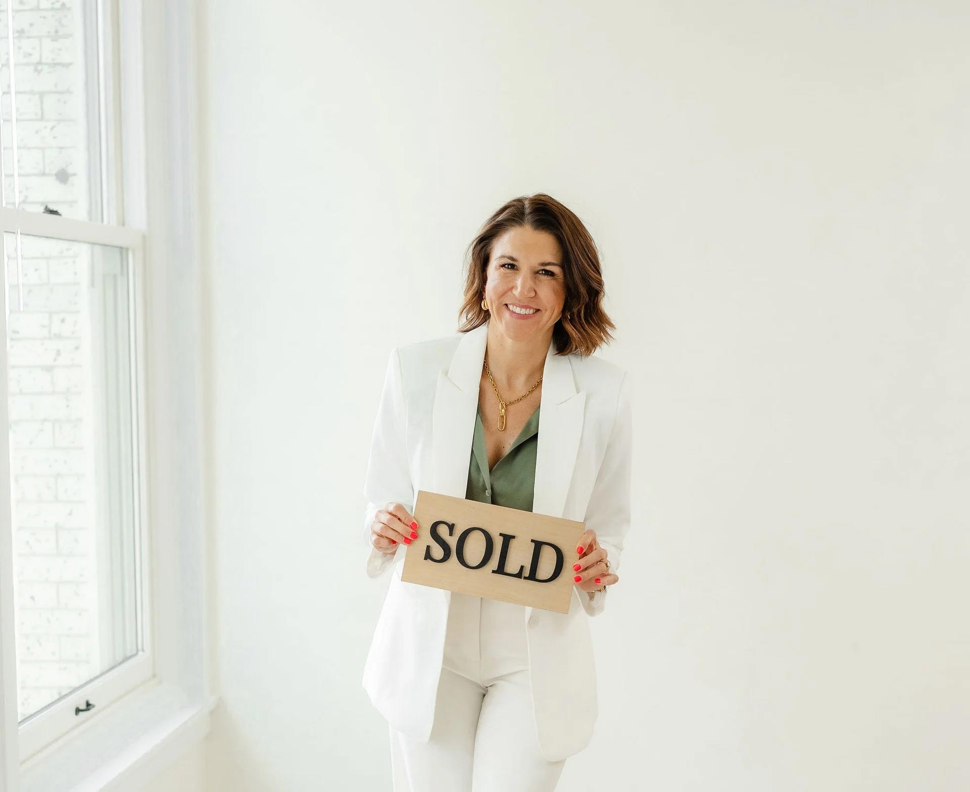 Real estate agent in white blazer holding a SOLD sign, smiling confidently